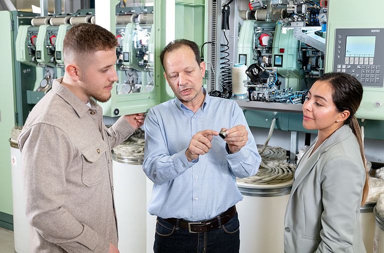 Three people stand in front of the control unit of a draw frame and discuss 
