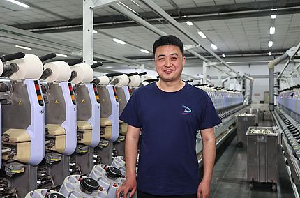 Zhang Sheng, Chief Engineer bei Sanyang Textile Co., Ltd., steht nach der Installation des Saugdüsen-Upgrades vor der Spulmaschine Autoconer 338.