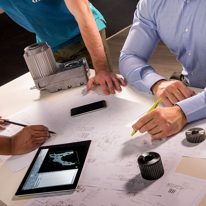 Three people standing at a table, machine parts and drawings in focus